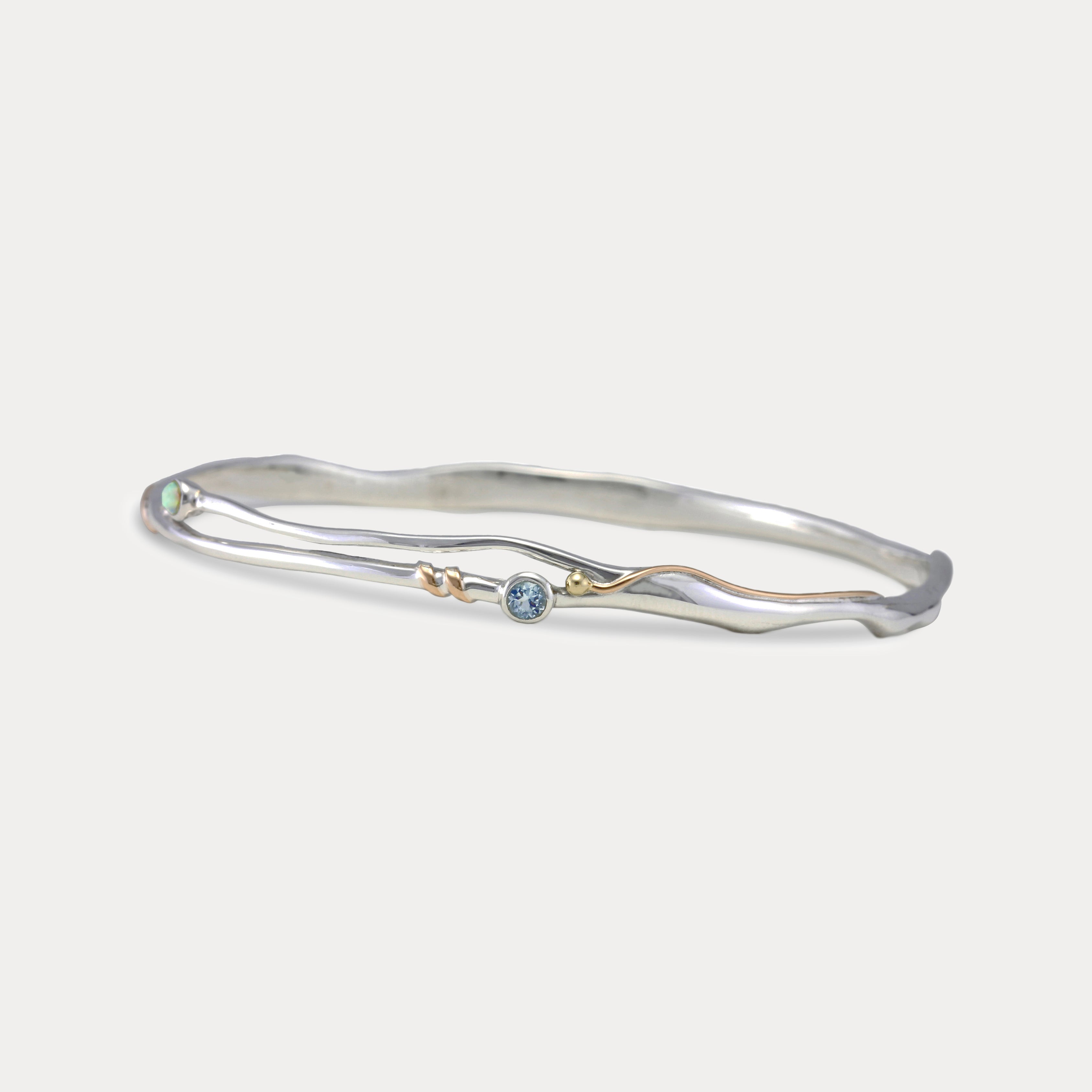 A Sterling Silver bangle bracelet with a small round blue Topaz and white Opal accented by 14KT Gold Fill Wire.