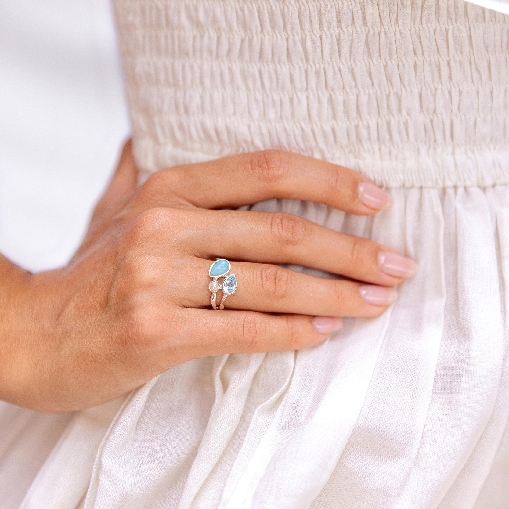 Close-up of a hand wearing a ring with a blue gemstone topaz and larimar and a white pearl against a white background