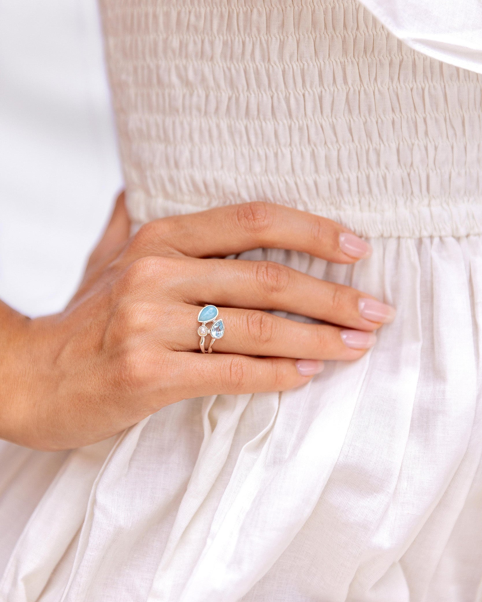 Close-up of a hand wearing a ring with a blue gemstone topaz and larimar and a white pearl against a white background